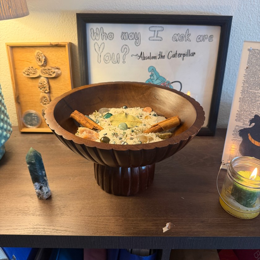 A wooden bowl on a pedestal holds rice, herbs, and crystals. It is flanked on the left by a moss agate crystal tower and on the right by a glass votive holder with a green candle that is lit.